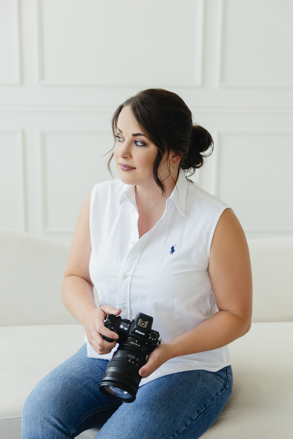woman holding a Nikon Z6ii camera in her hands while sitting on a white couch in a white wall photo studio looking out the window